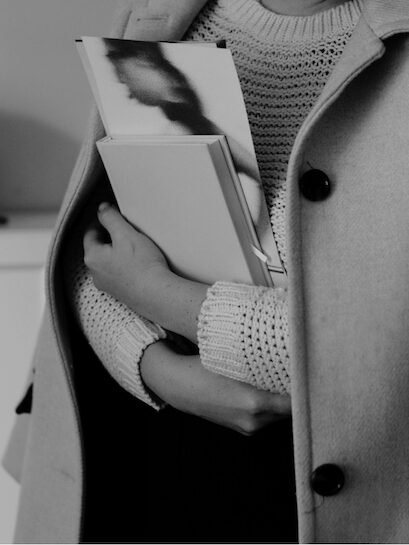 A female solopreneur in a knitted sweater and buttoned coat clutches email list books to her chest.