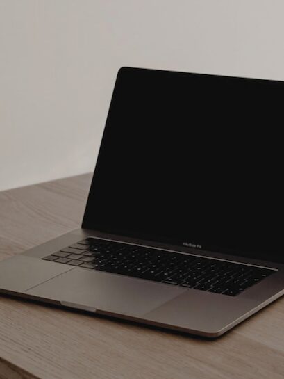 A female solopreneur's MacBook Pro sits open on a wooden desk, its black screen off and keyboard visible against a plain wall. She sets up her email list.