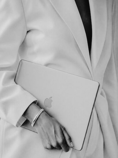 A female entrepreneur wearing a white blazer and trousers is walking while holding a MacBook, on which she has recently started building her email list.
